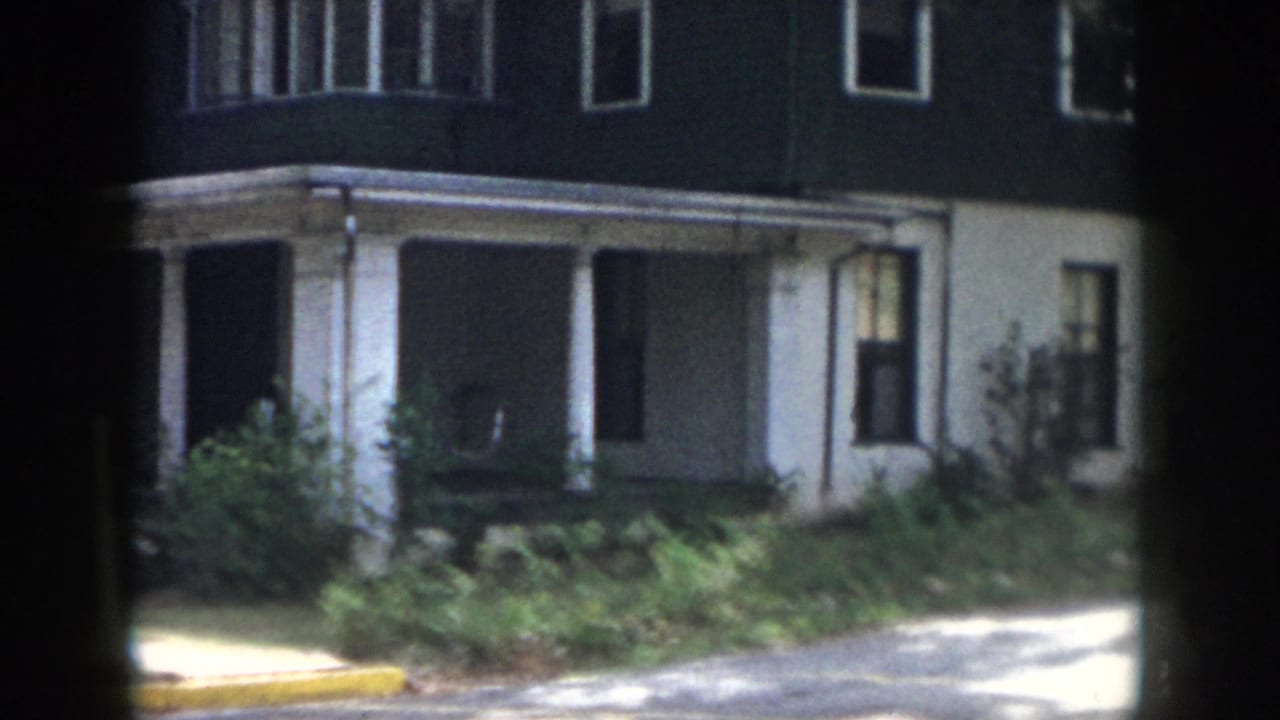 1958 Dayton exterior view of the house corner, porch columns, dark upper siding, white lower wall, shrubs, and sunlit pavement.