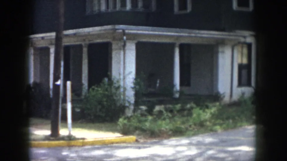 1958 Dayton frame showing the house porch from the street with a utility pole, yellow curb, shrubs, sidewalk, and shaded facade.