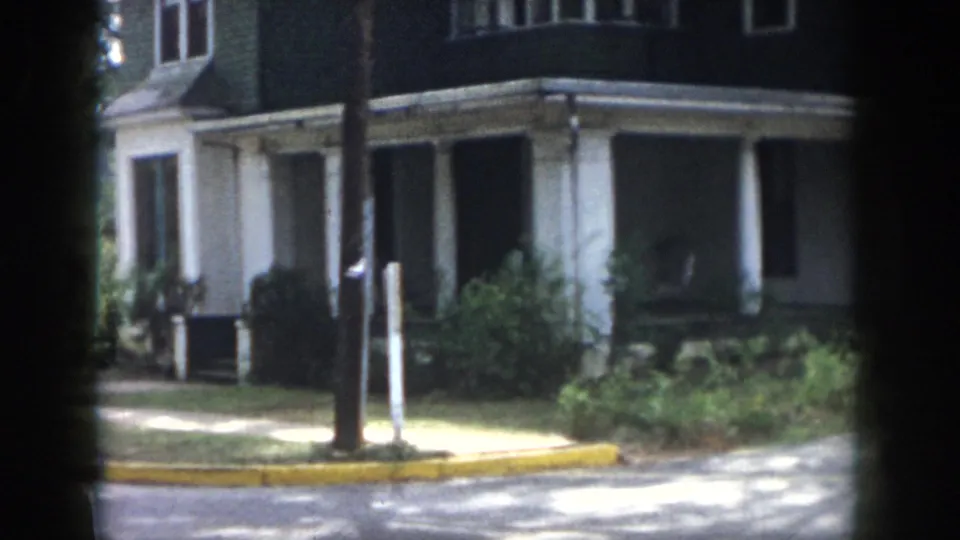 1958 Dayton home-movie frame of the street-corner house with porch posts, shrubs, yellow curb, sidewalk, and dappled sunlight.