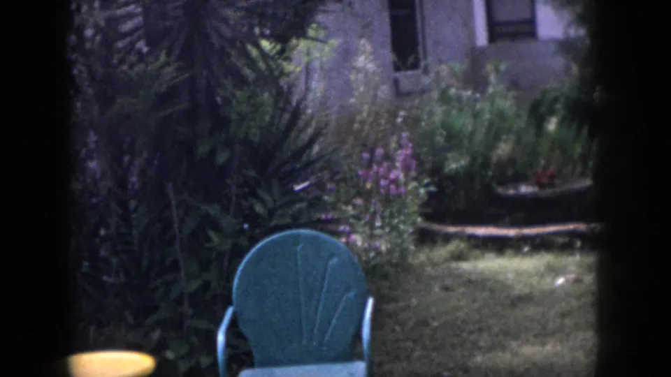 1958 Dayton backyard view with a teal chair, dense planting, stone-edged garden feature, lawn, and partial house exterior.