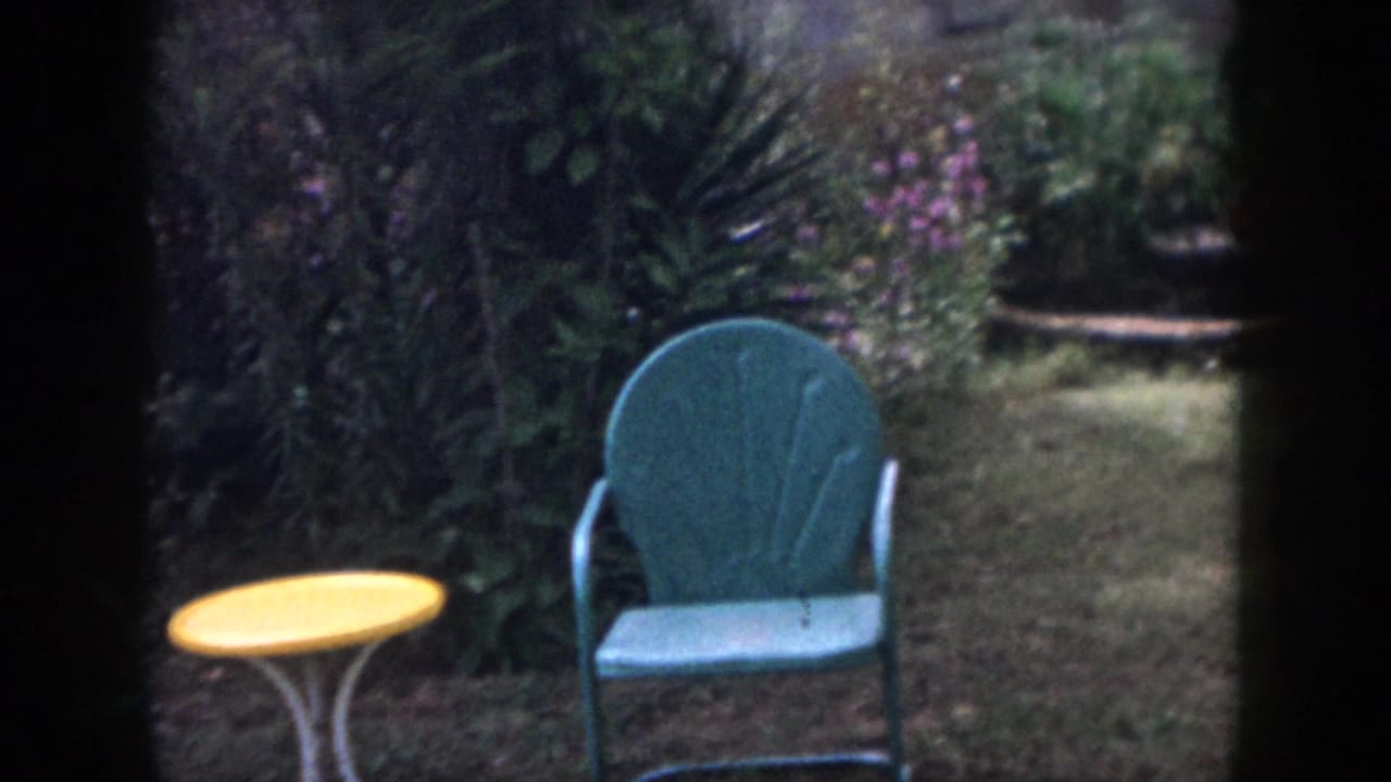 1958 Dayton home-movie frame showing a teal patio chair, yellow table, grass, and dense garden planting seen through dark framing.
