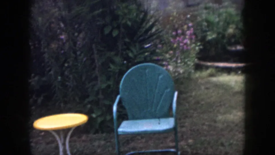1958 Dayton home-movie frame showing a teal patio chair, yellow table, grass, and dense garden planting seen through dark framing.