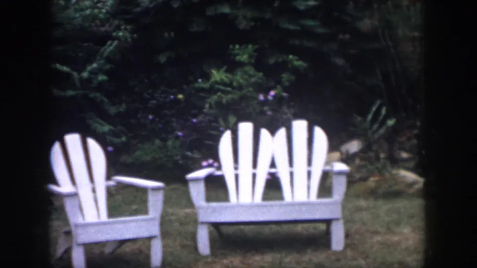 1958 home-movie frame of a white Adirondack-style chair and matching bench on a backyard lawn in Dayton, Ohio.