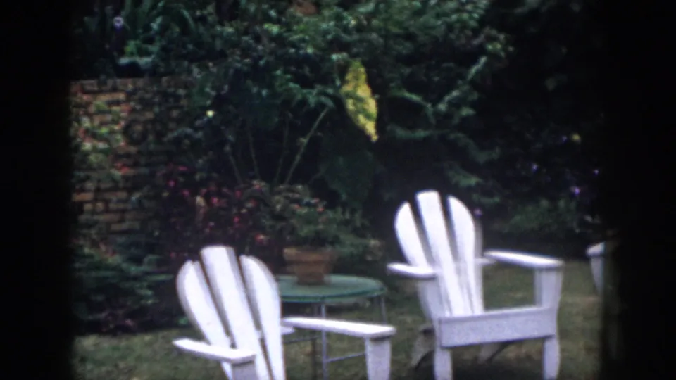 1958 Dayton home-movie frame showing white outdoor chairs in a leafy garden beside a brick wall.