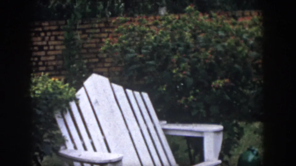 1958 Dayton home-movie frame with a white Adirondack-style chair in front of shrubs and a brick garden wall.