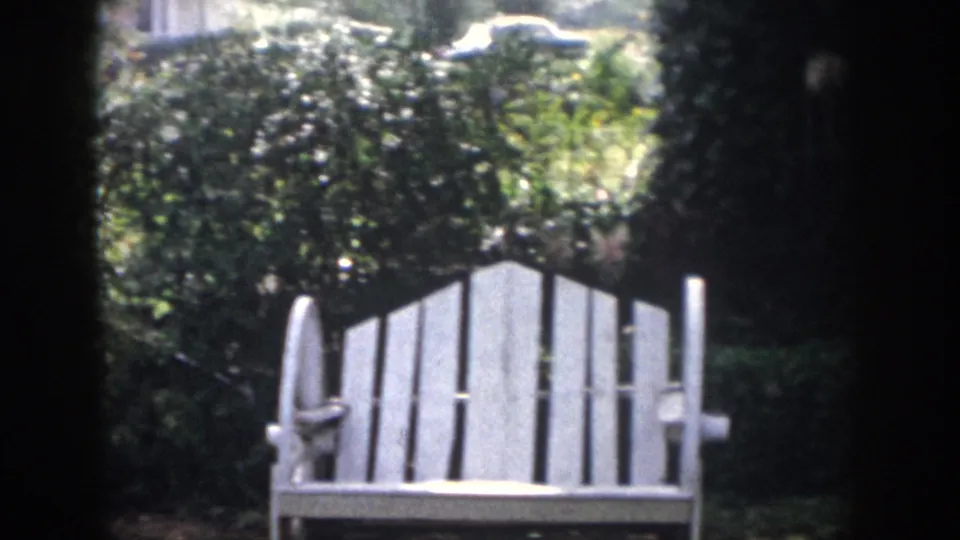 1958 Dayton, Ohio home-movie frame with a white Adirondack-style chair set among shrubs, vines, and strong midday garden light.