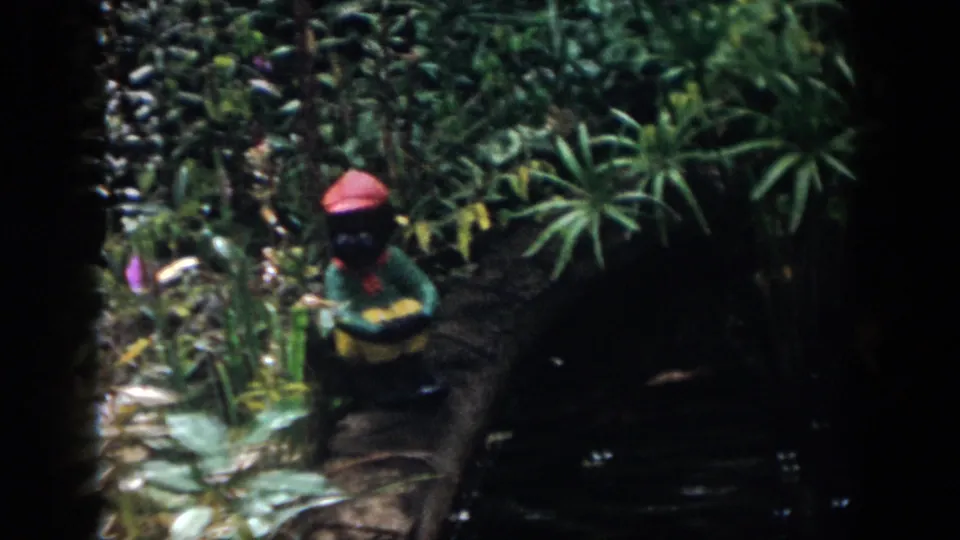 1958 Dayton home-movie frame showing a red-hatted garden gnome near pond plants, water lilies, and shaded water.