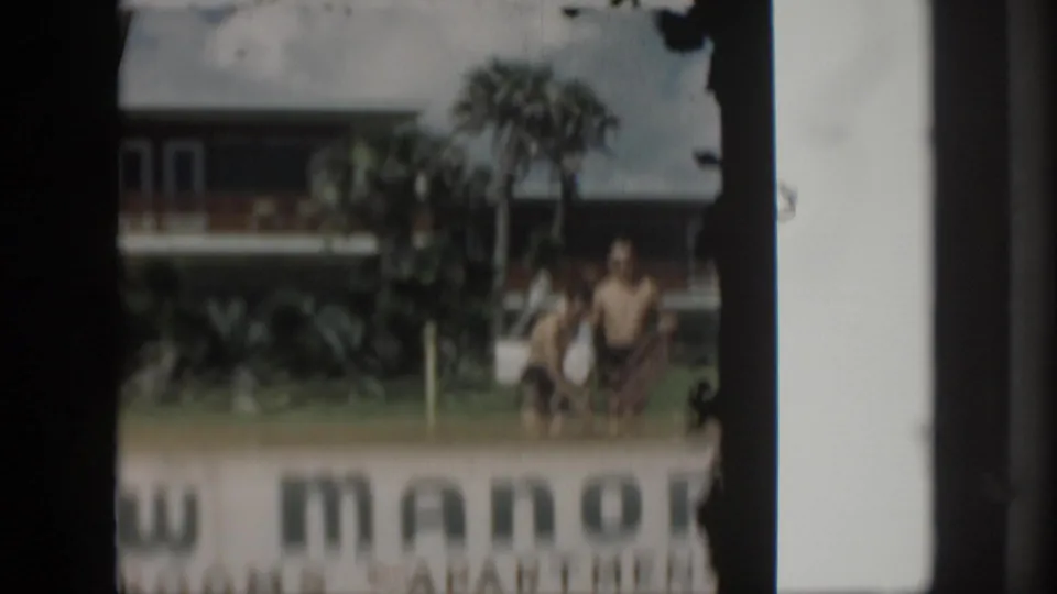 1958 home-movie frame with a weathered apartment-room sign, lawn, palms, and two swimwear-clad figures in the background.