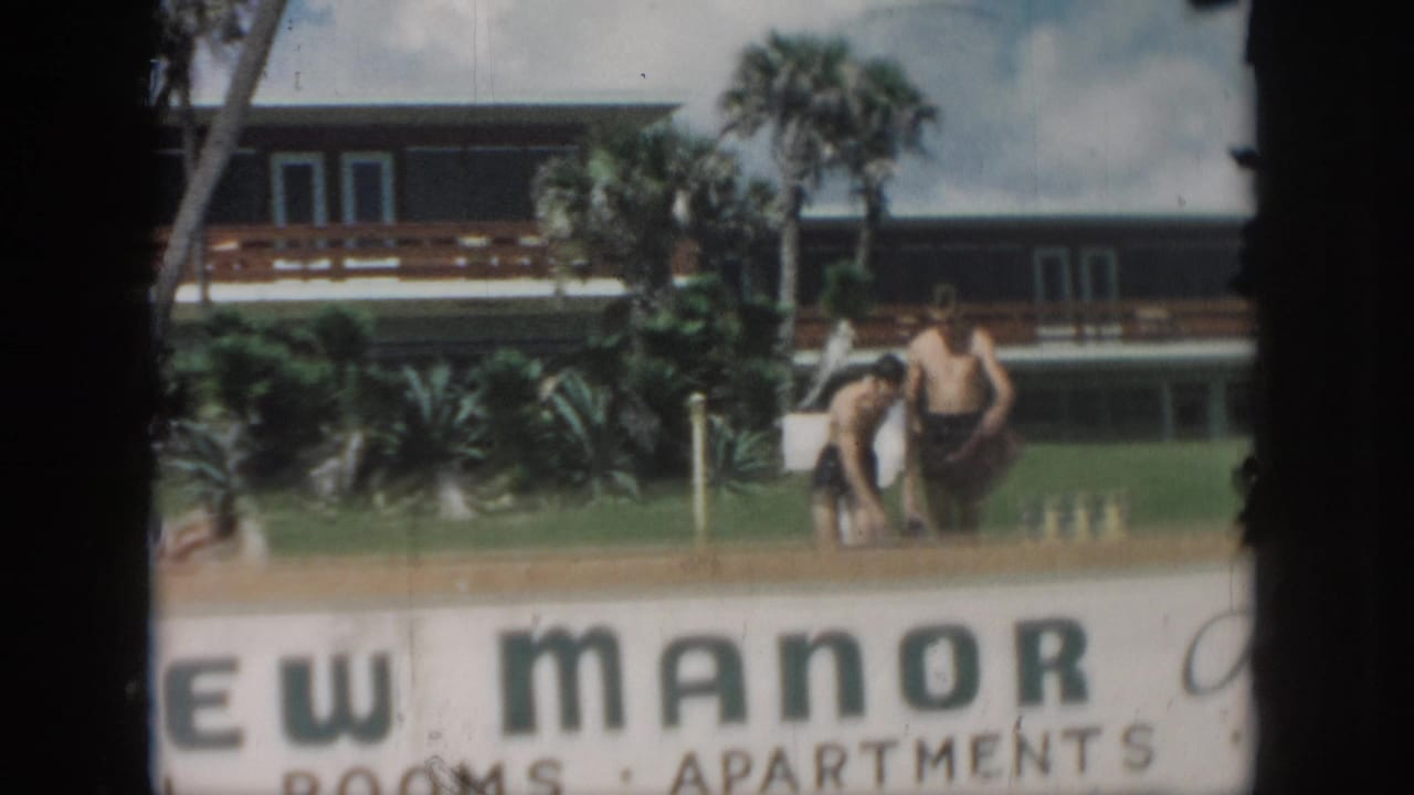1958 New Orleans frame with two men in swim trunks near the View Manor sign, palm landscaping, and hotel-apartment building.