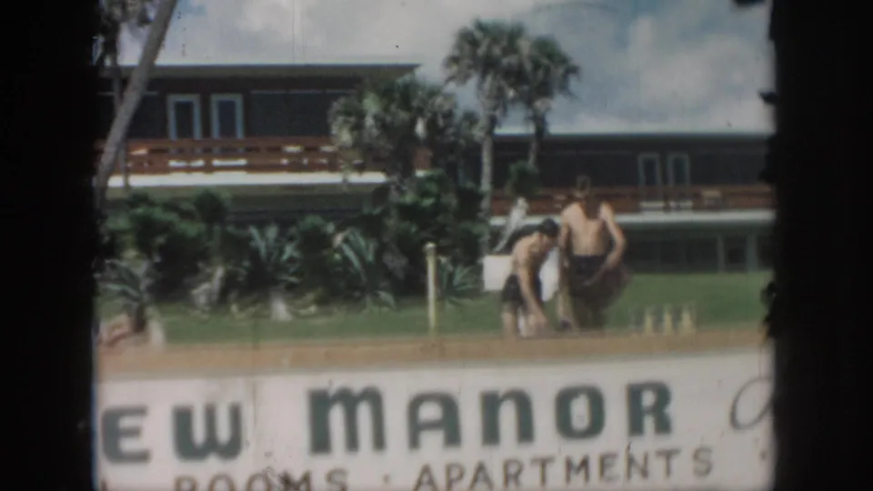 1958 New Orleans frame with two men in swim trunks near the View Manor sign, palm landscaping, and hotel-apartment building.