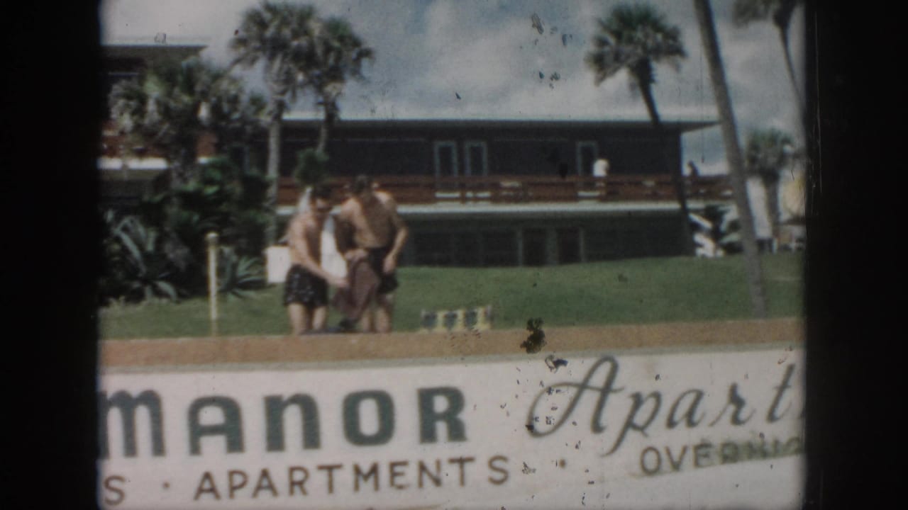 Two young men stand on a lawn behind a Manor Apartments sign with palm trees and an apartment building behind them.