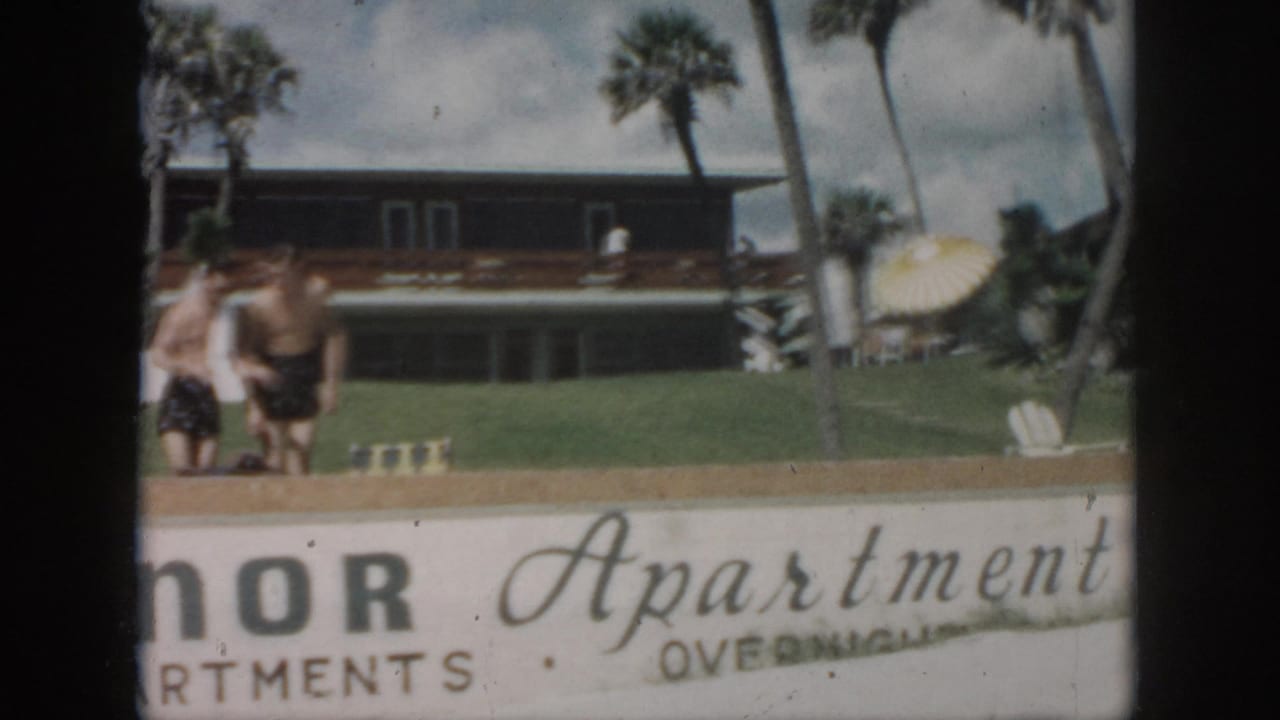 Two young men remain near the lawn as the Manor Apartments sign fills the foreground and palm trees rise behind them.