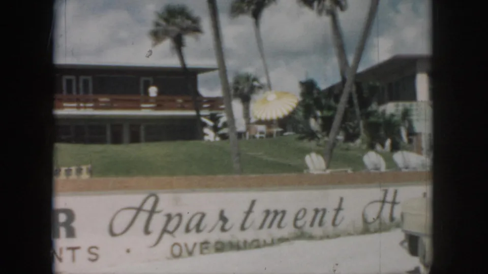 1958 New Orleans home-movie view of the apartment lawn, palms, outdoor chairs, and sun umbrella behind the cropped Manor Apartments sign.