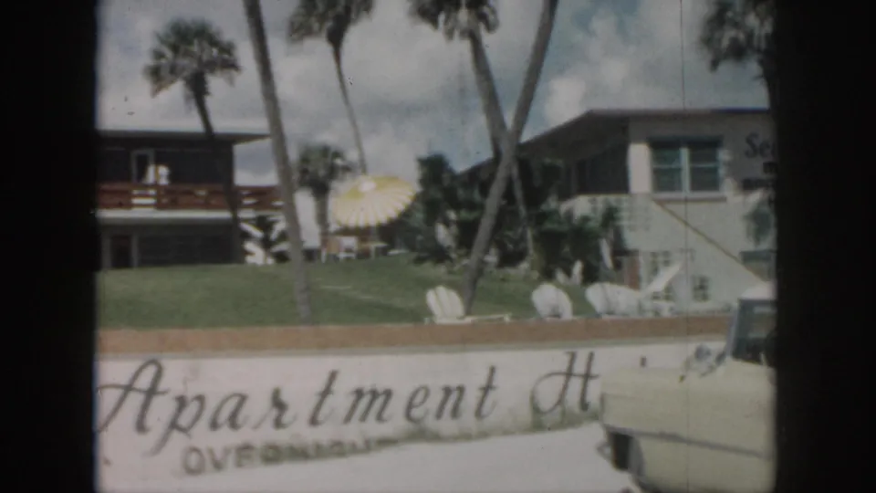 1958 New Orleans frame showing the apartment grounds with palms, lawn seating, a sun umbrella, and the cropped Manor Apartments sign.