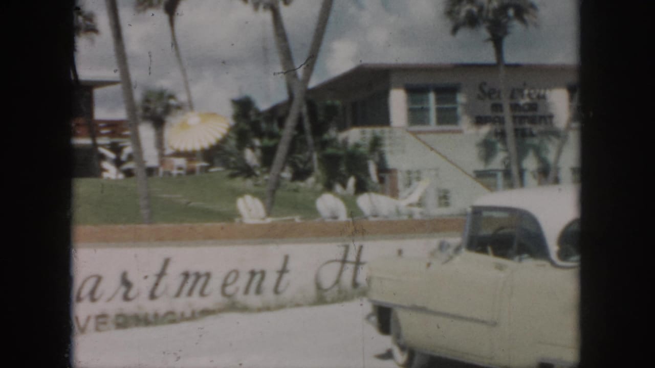 1958 New Orleans home-movie frame showing a cream classic car in front of Sea View Motor Inn Apartment Hotel signage and palms.