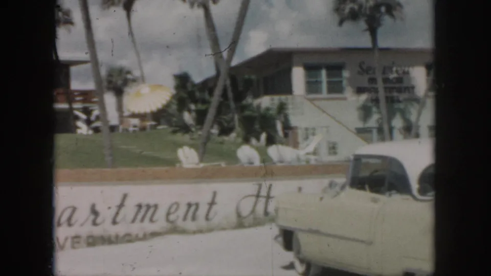 1958 New Orleans home-movie frame showing a cream classic car in front of Sea View Motor Inn Apartment Hotel signage and palms.