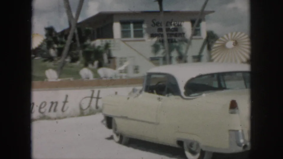 1958 New Orleans frame of a cream classic car parked by the Sea View Motor Inn exterior with palms and sunburst decor.