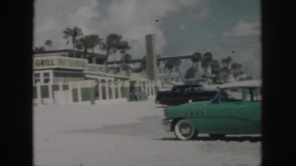 1958 New Orleans roadside frame showing Spray Grill frontage, a green mid-century car, a black parked car, and palm trees behind the building.