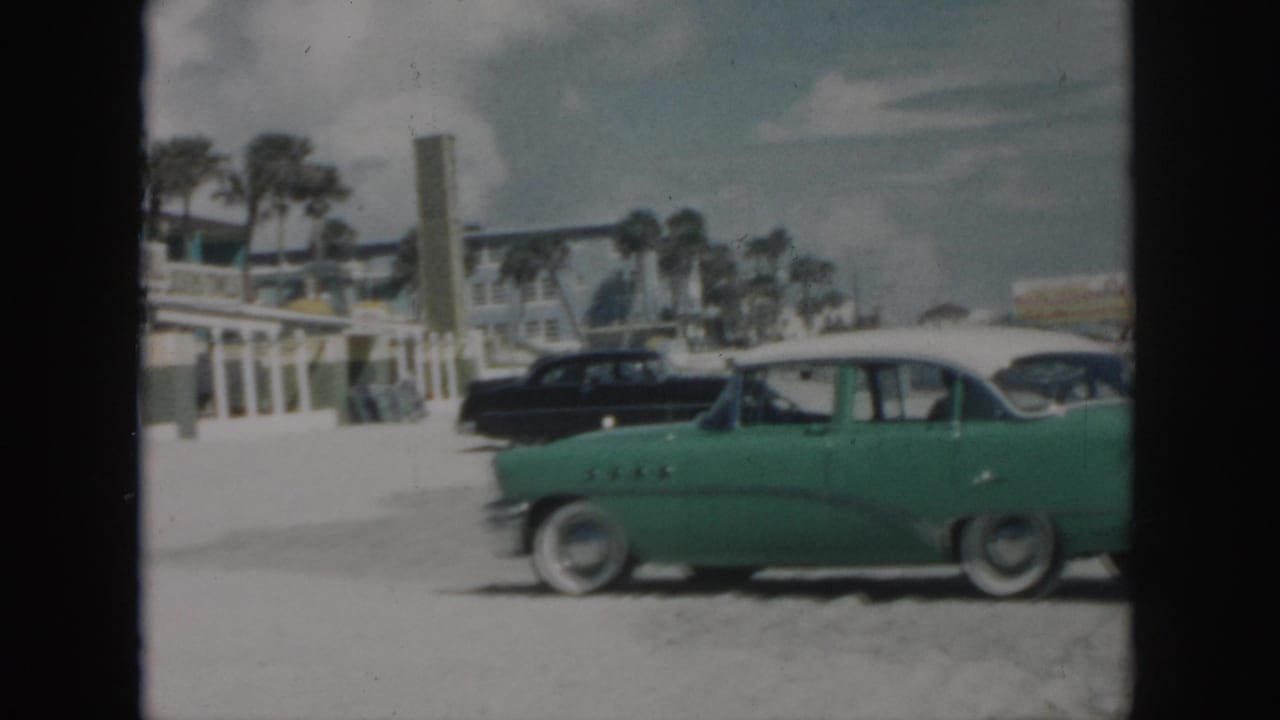Green classic car with white roof parked on sandy beach near palm trees and buildings.