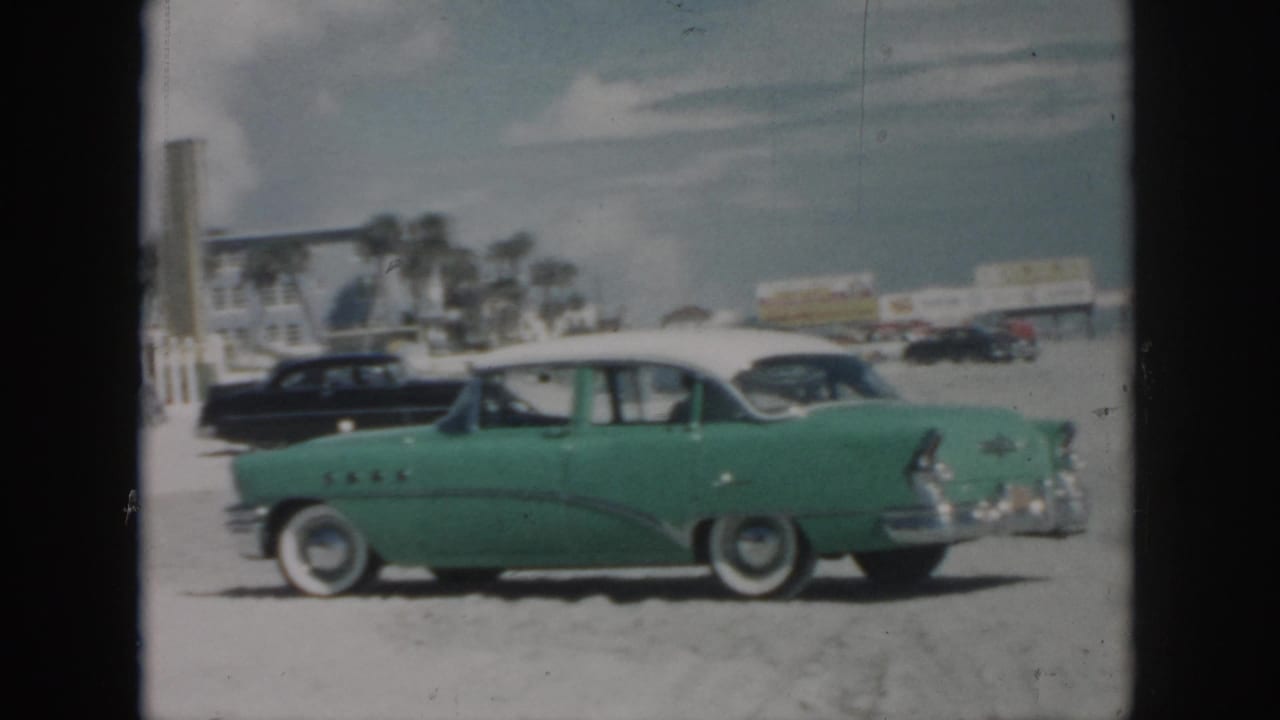 Side view of green classic car parked on sand with black sedan and beach buildings behind it.