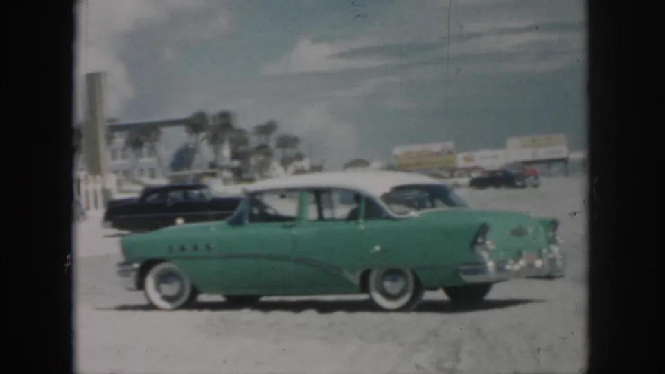 1958 New Orleans beachside frame with a green white-roof classic car, black sedan, palm trees, and low commercial buildings in the background.