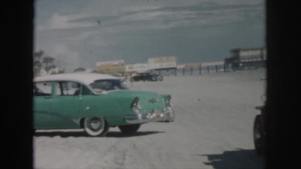 1958 New Orleans home-movie view of the green classic car from the rear side, with beachside buildings and parked cars beyond.