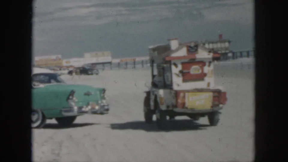1958 New Orleans beachside frame showing the green classic car near a small food cart, with low buildings and parked cars in the background.