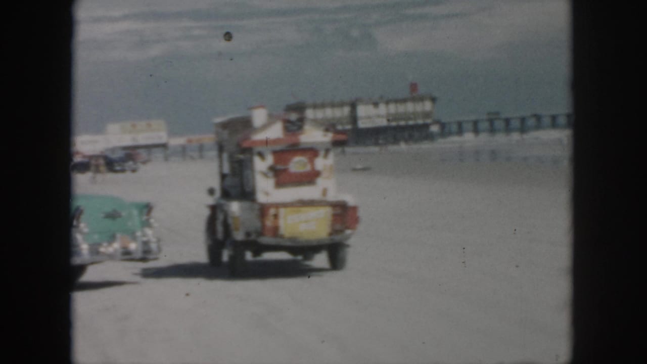 1958 New Orleans beach footage showing a turquoise car, a small truck, sand, surf, and a long pier in the background.