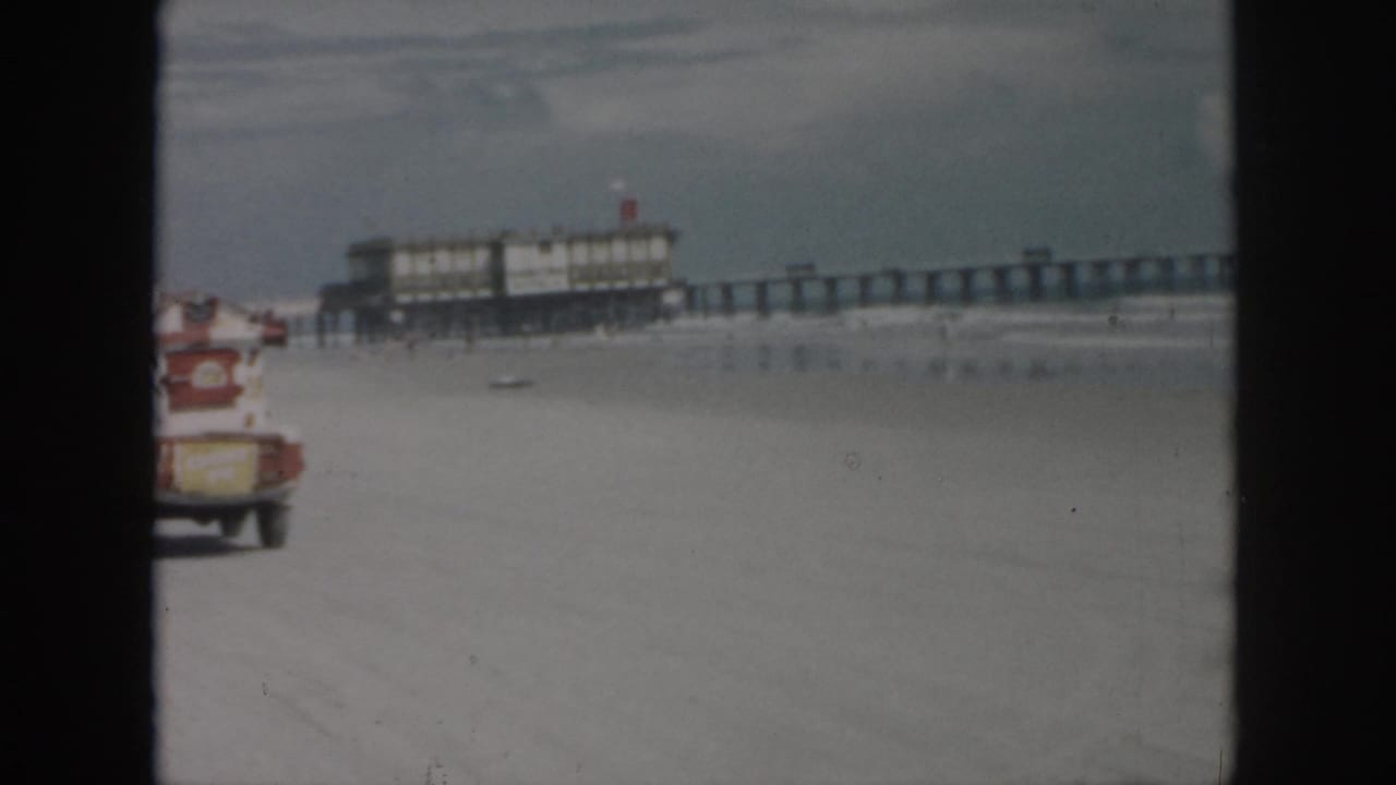 Small truck drives across a sandy beach with a pier behind it.