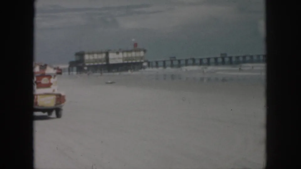 1958 New Orleans beach scene with a small truck moving away on sand and a long pier stretching over the water.