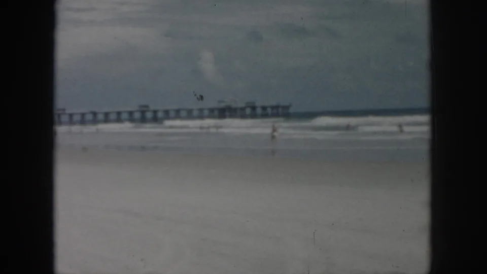 Wide 1958 beach view in New Orleans with muted 8mm color, a pier, gentle surf, and a distant beachgoer.