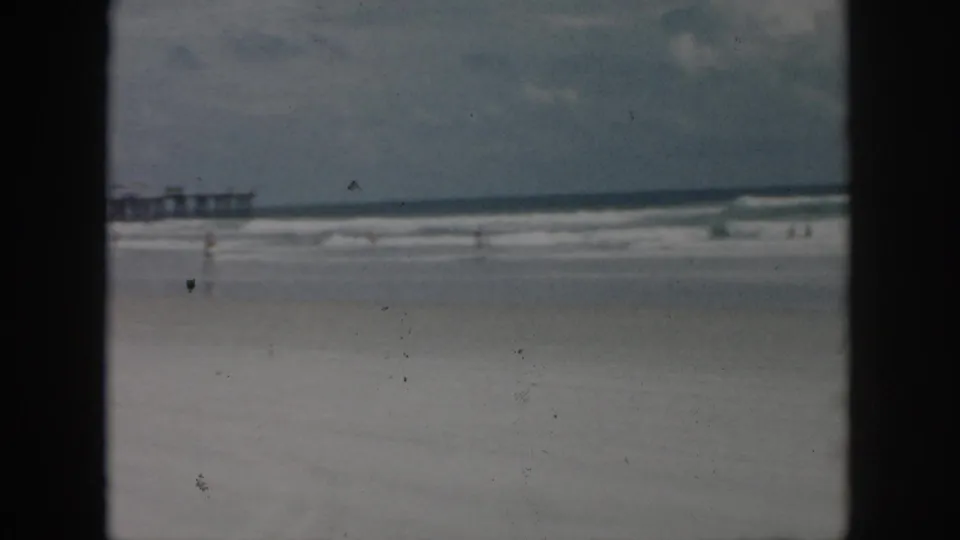 1958 New Orleans beach frame with rolling surf, wet sand, partial pier, and distant people near the shoreline.