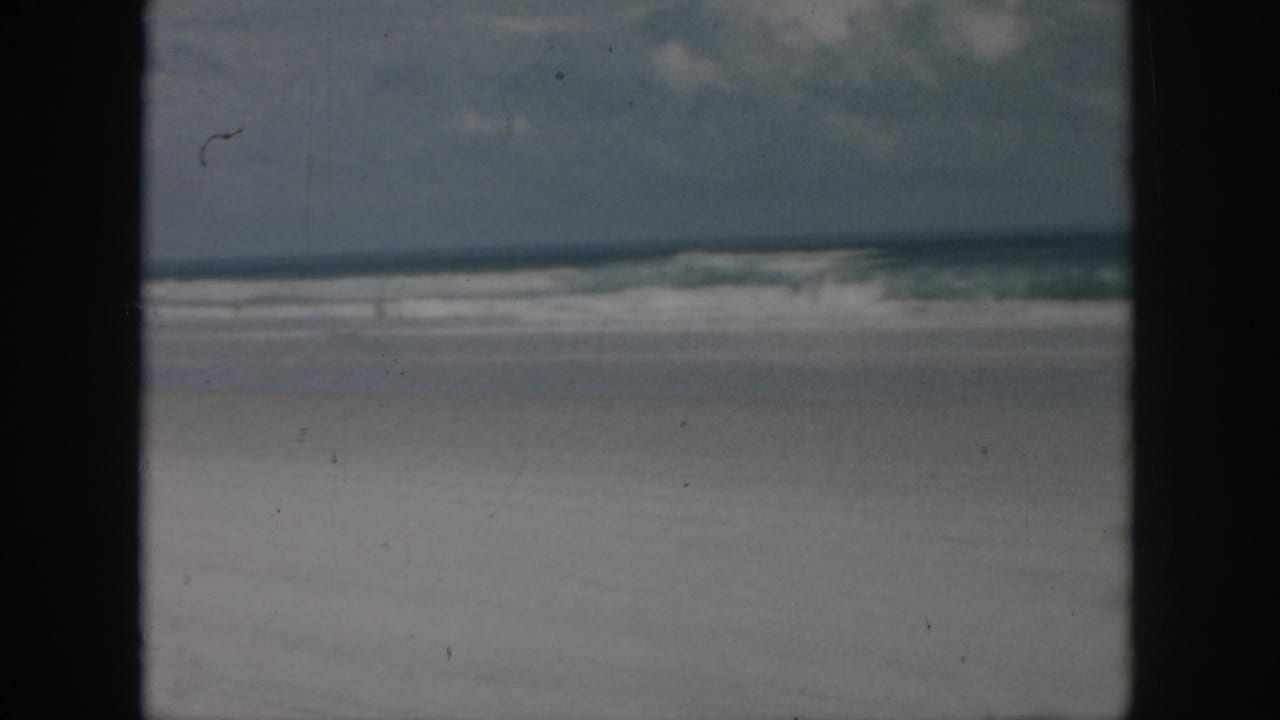 1958 8mm home-movie frame from the New Orleans, Louisiana metadata showing an empty sandy shoreline, breaking waves, cloudy sky, and dark foreground framing.