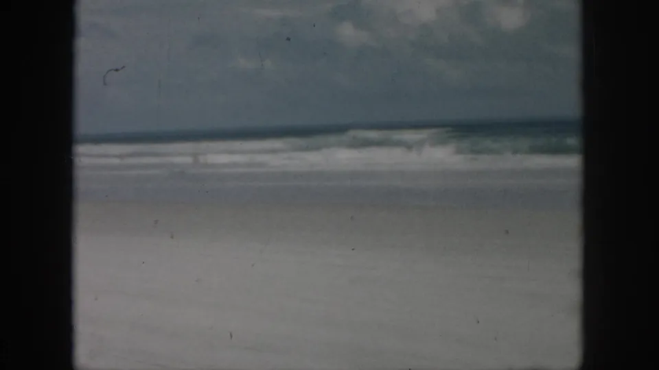1958 8mm home-movie frame from the New Orleans, Louisiana metadata showing an empty sandy shoreline, breaking waves, cloudy sky, and dark foreground framing.