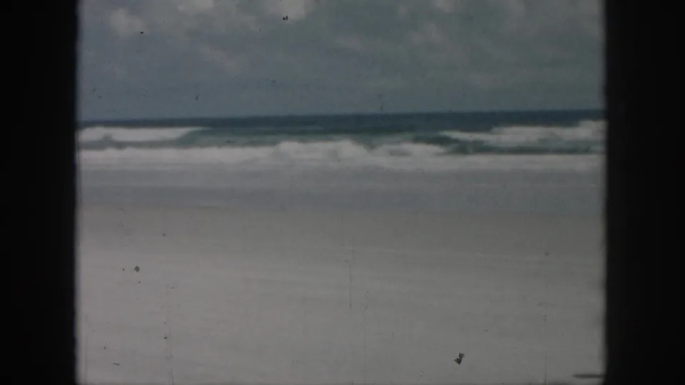 Wide 8mm beach view with pale sand, rolling surf, cloudy sky, and dark vertical edges that partially obscure the shoreline.