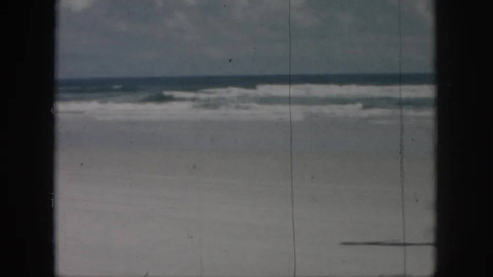 1958 home-movie beach frame showing surf and open sand, with a partial dark form near the lower right edge as the action moves into view.