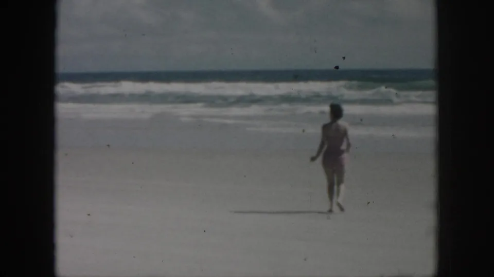 1958 New Orleans, Louisiana 8mm frame showing a woman in a bikini running across the beach toward breaking ocean waves under a cloudy sky.