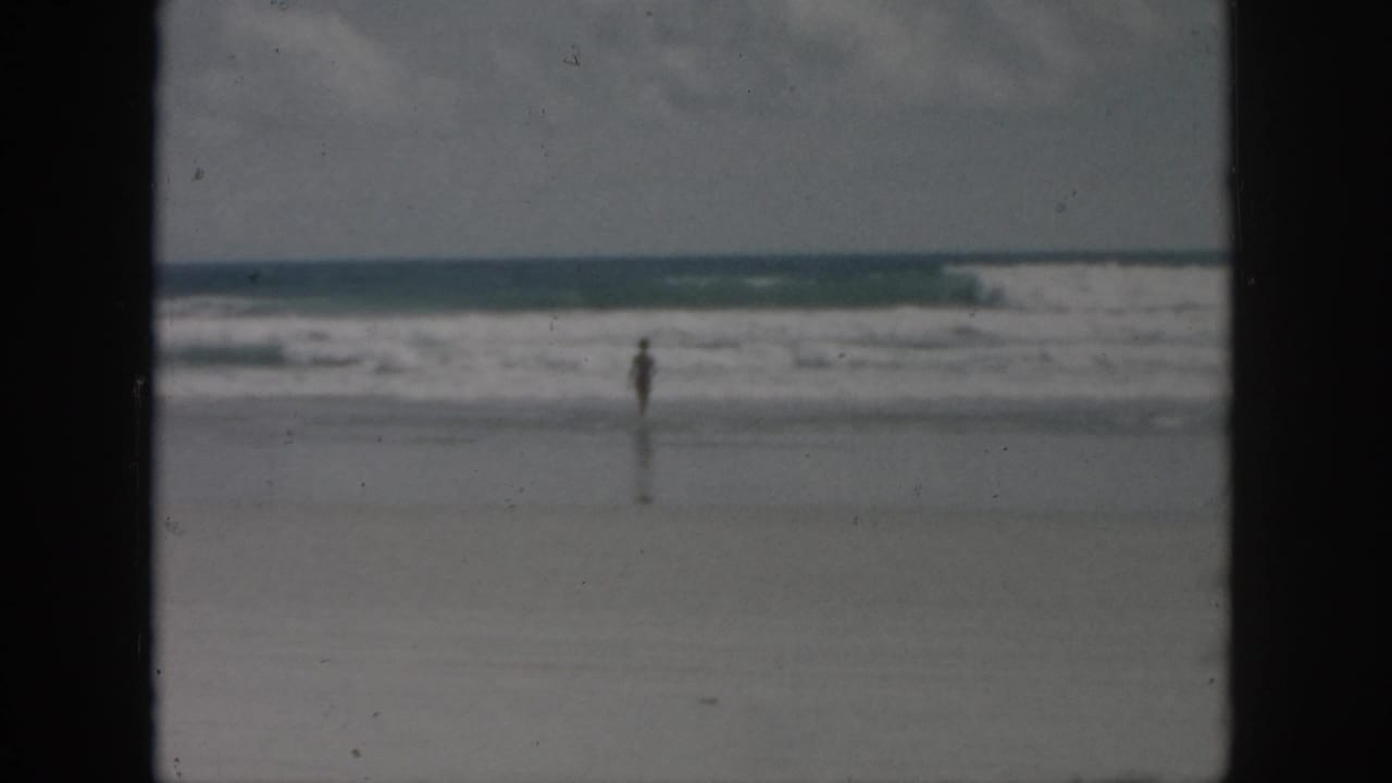 1958 8mm home-movie frame of a distant person near the surf on a sandy beach, with soft focus and dark film borders.