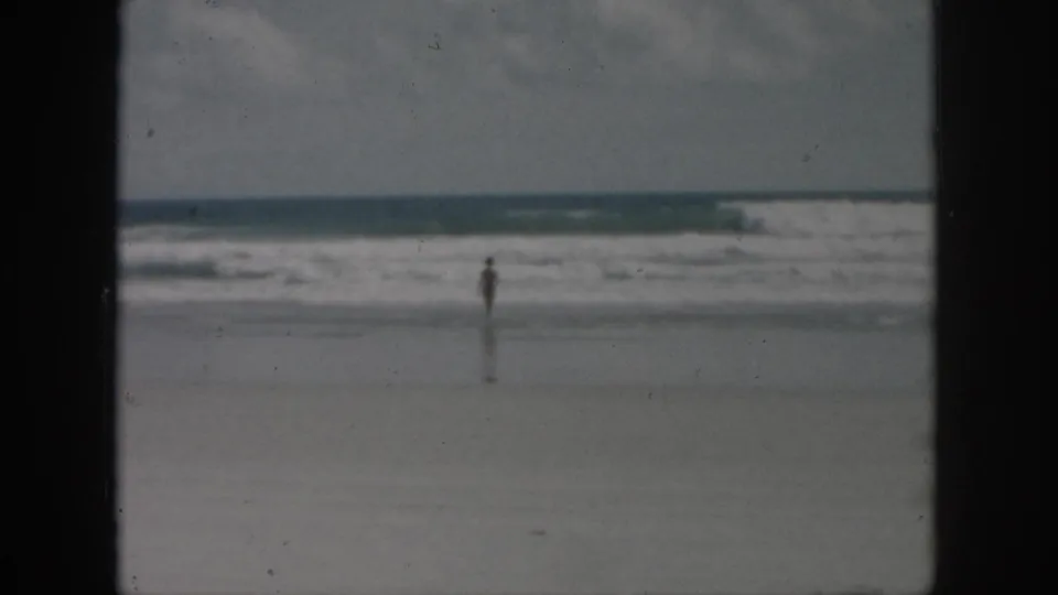 1958 8mm home-movie frame of a distant person near the surf on a sandy beach, with soft focus and dark film borders.