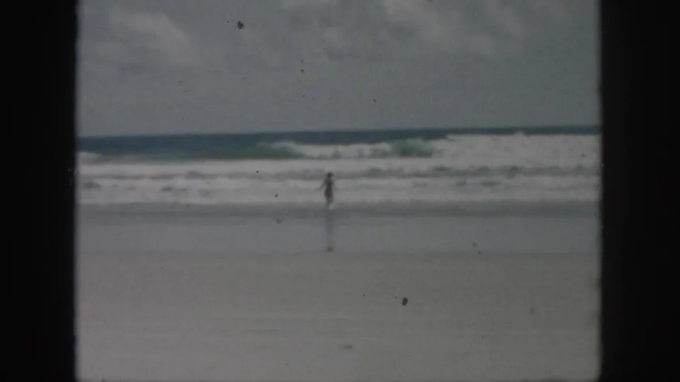 Distant figure on a beach in 1958 home-movie footage, framed by surf, wet sand, muted color, and 8mm gate edges.