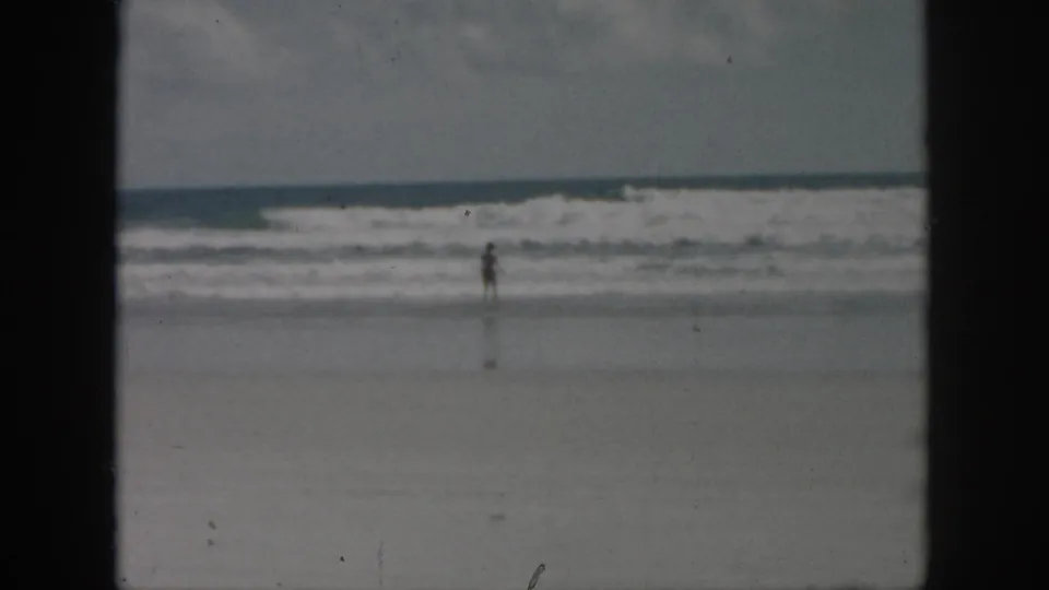 1958 8mm frame showing a distant beachgoer at the surf line, with rolling waves and a soft, faded home-movie look.
