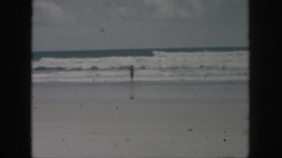 Final 1958 8mm beach frame with a small solitary figure near the waves, muted color, soft focus, and visible film wear.