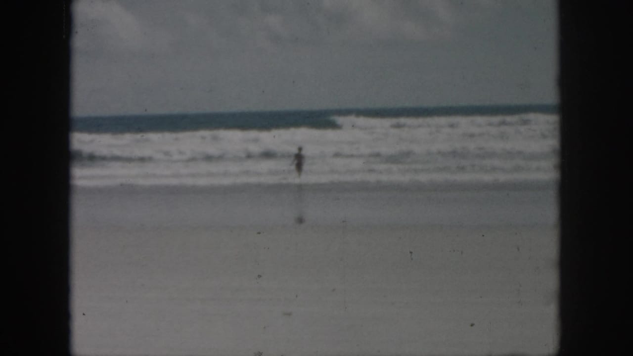 1958 8mm home-movie frame from New Orleans metadata showing a distant figure in shallow ocean water beneath an overcast sky.