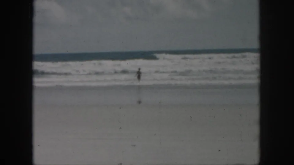 1958 8mm home-movie frame from New Orleans metadata showing a distant figure in shallow ocean water beneath an overcast sky.
