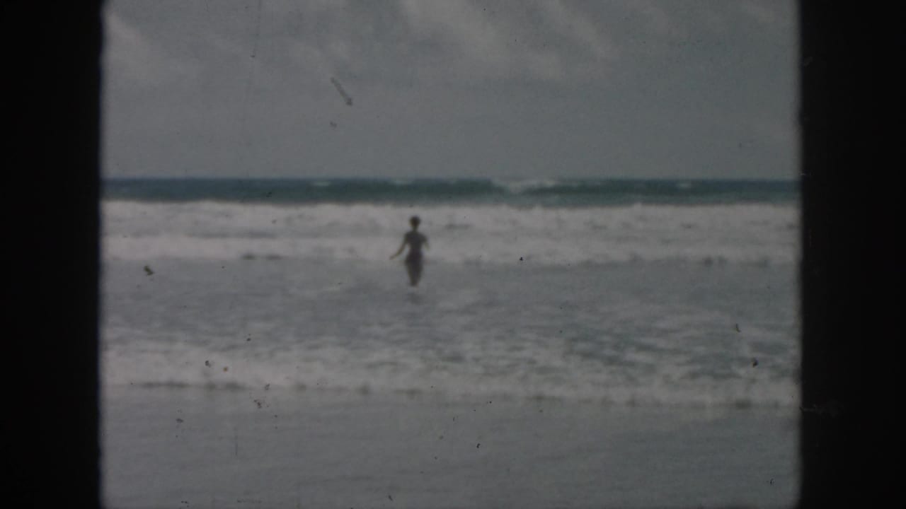 Distant figure wades through shallow beach water in a soft, grainy 1958 8mm frame associated with New Orleans, Louisiana.
