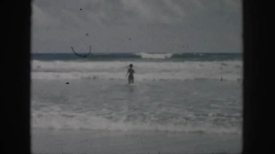 A distant person stands or walks in shallow ocean water, framed by visible 8mm borders and muted late-1950s color.
