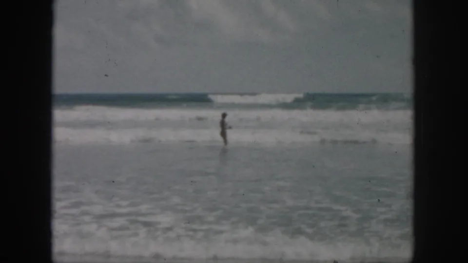 Soft 8mm frame shows a small figure in profile at the shoreline, with rolling ocean waves under a gray sky.