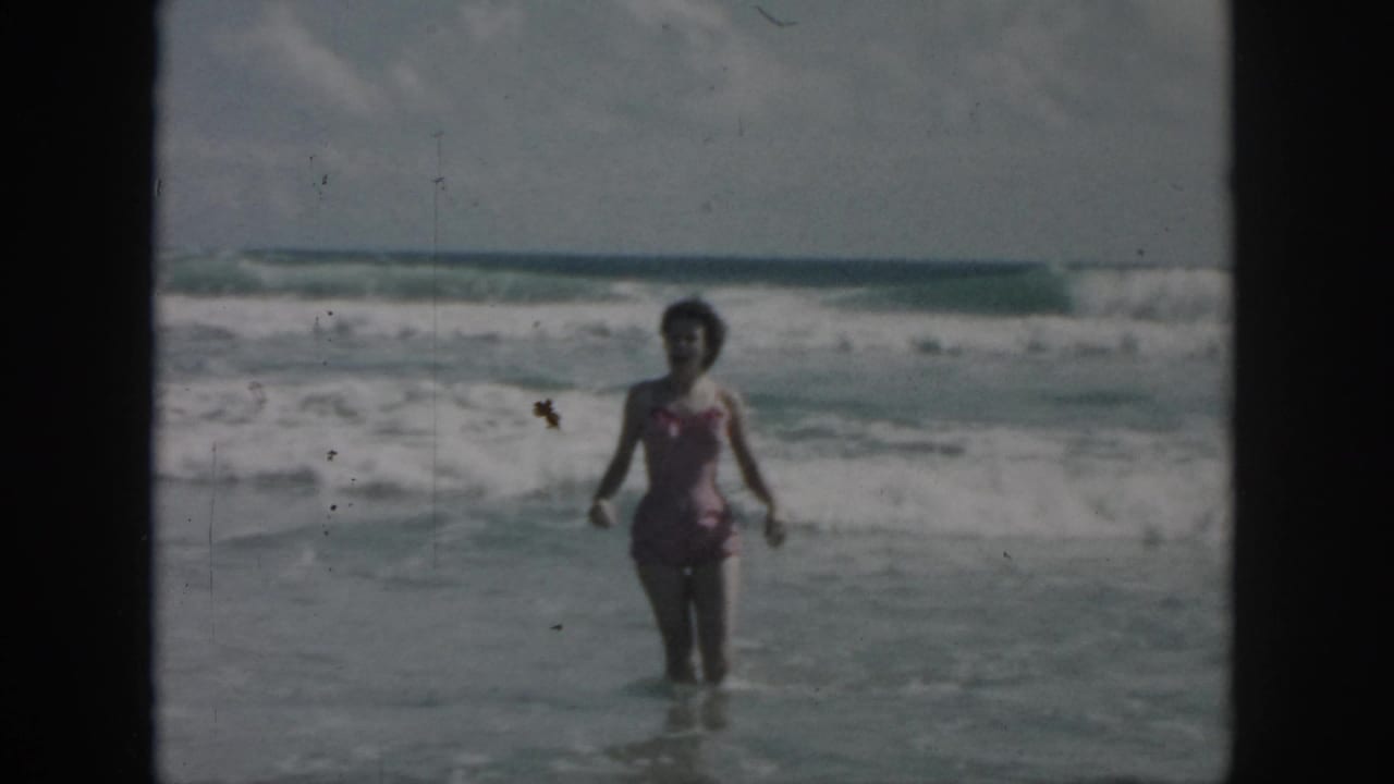1958 home-movie frame of a woman in a pink one-piece swimsuit wading in choppy surf, with black 8mm borders visible.