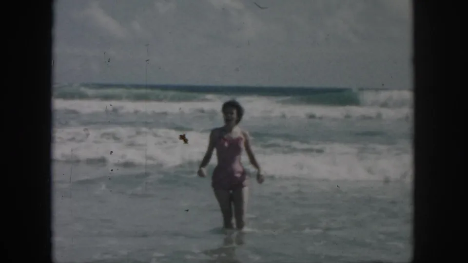 1958 home-movie frame of a woman in a pink one-piece swimsuit wading in choppy surf, with black 8mm borders visible.