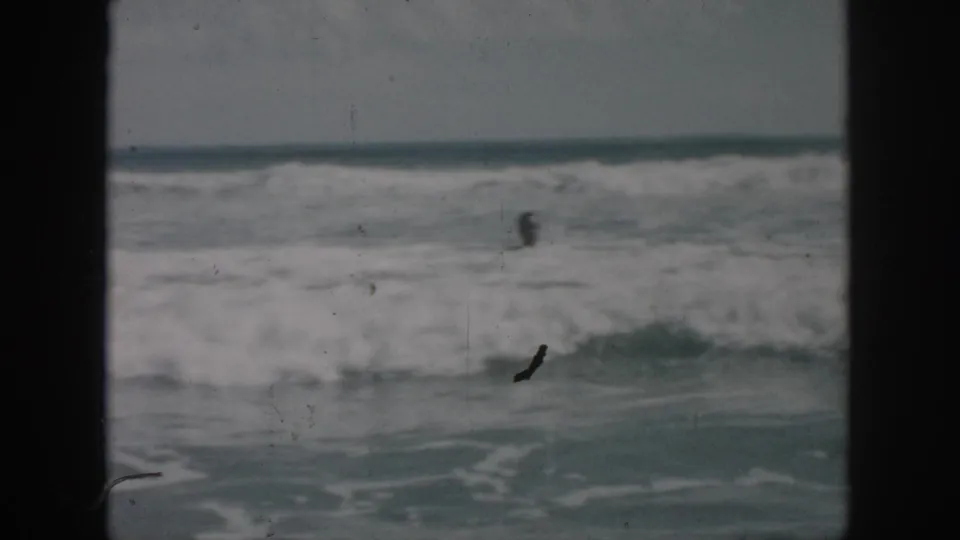 1958 home-movie view of rolling surf and a distant swimmer, framed by dark 8mm film borders.